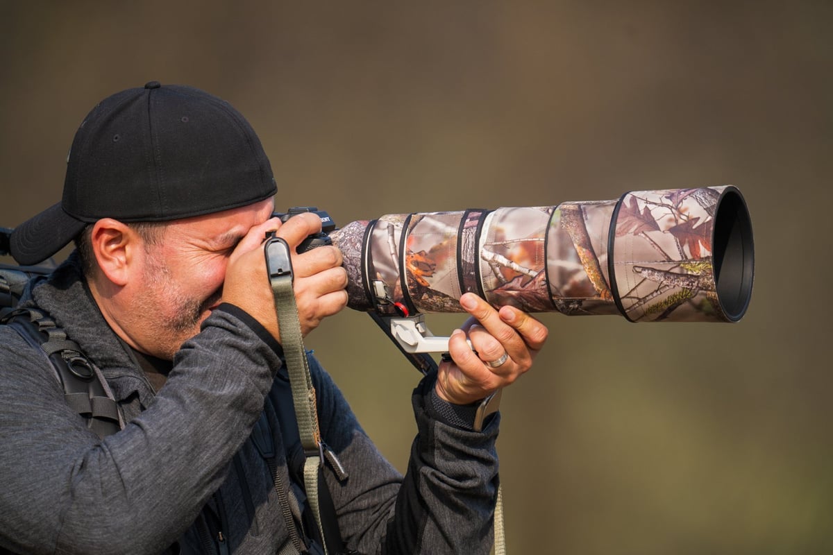 Photographer wearing a cap holding a camouflaged telephoto lens and camera against a blurred brown background
