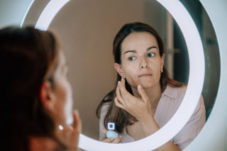 Woman examining her face in a lit vanity mirror with her hand to her chin