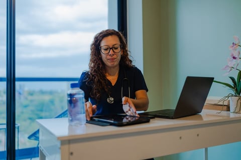 Female healthcare worker wearing glasses and stethoscope sits at desk with laptop in modern office overlooking cityscape