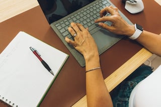 Persons hands typing on laptop keyboard at desk with notebook, pen, and wireless mouse
