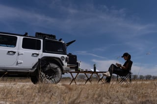 Person sitting in a camping chair next to a white lifted SUV in a dry grass field under blue sky